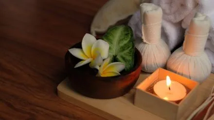 Wooden tray holding a small bowl with white-yellow frangipani flowers and a green leaf, two herbal compresses, and a lit tealight candle.