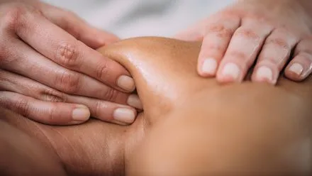 Massage therapist's hands kneading a person's shoulder and upper back, fingers pressing into the skin.